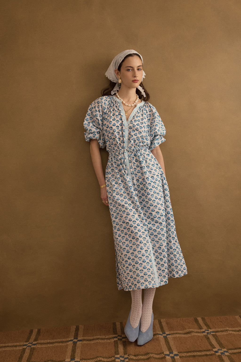 Editorial front view of model wearing the Juliet Block Printed Cotton Drop Sleeve Raglan Shirt Dress in Blue Gardenia, leaning against an olive green wall with a vintage organza scalloped headscarf