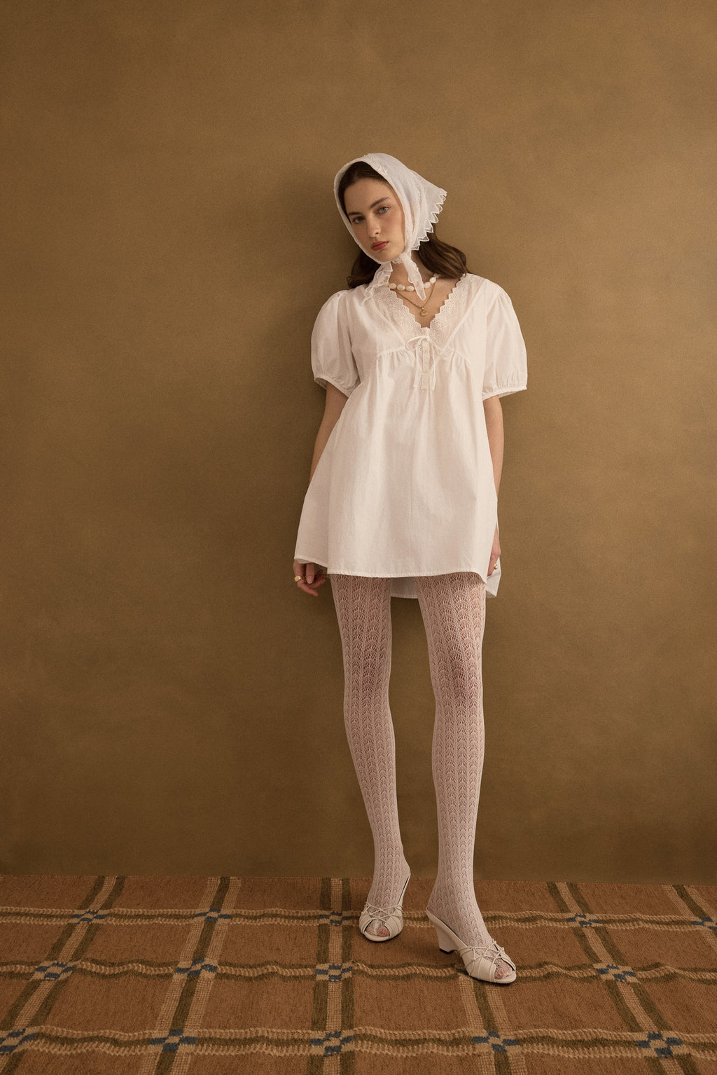 Model wears the Amelie organic cotton v-lace short dress in ivory, and stands styled against an olive green wall.
