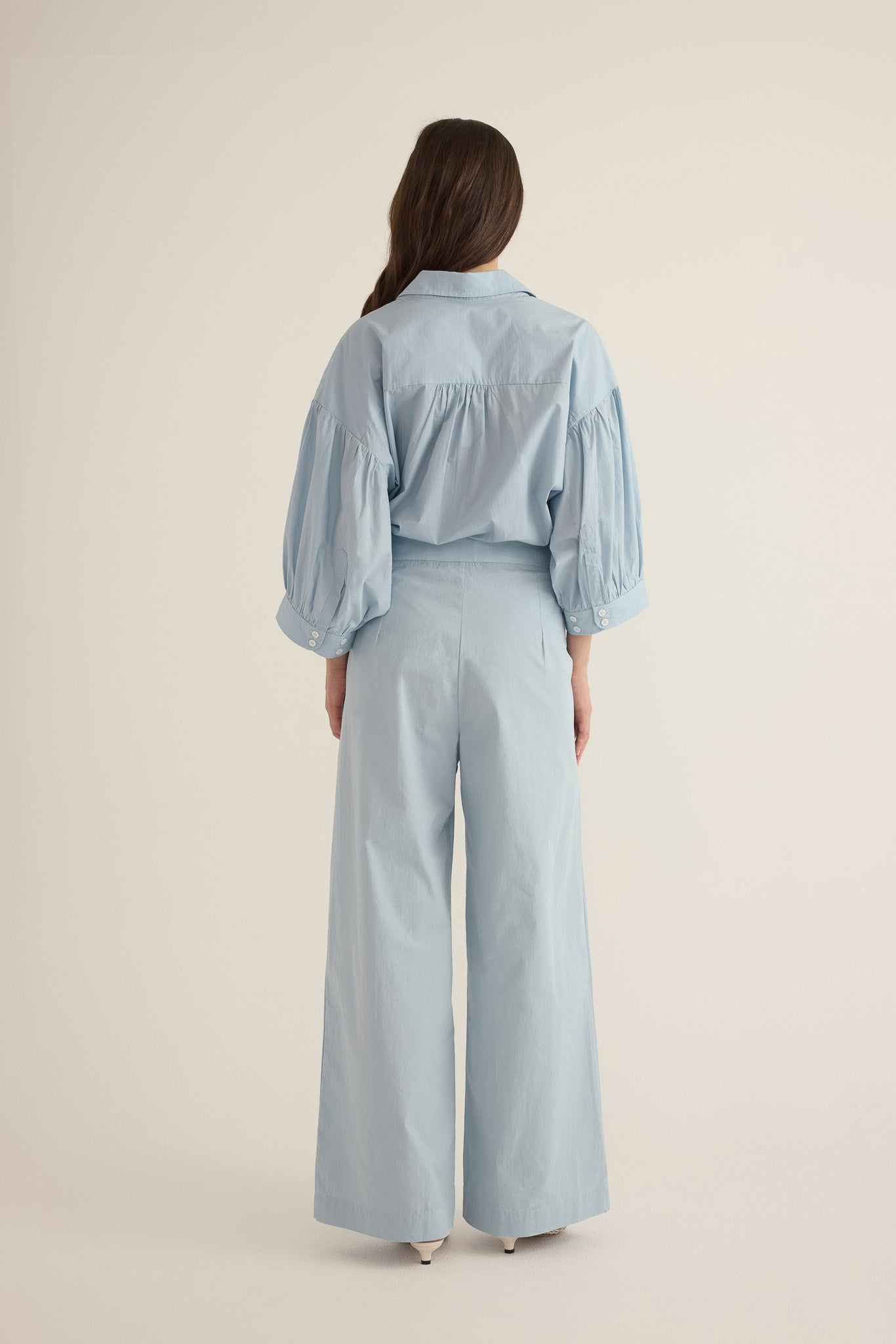 Back view of model wearing the Romeo wide-leg pleat pant in pale blue organic cotton, styled with the Romeo wide cuff blouson shirt in blue