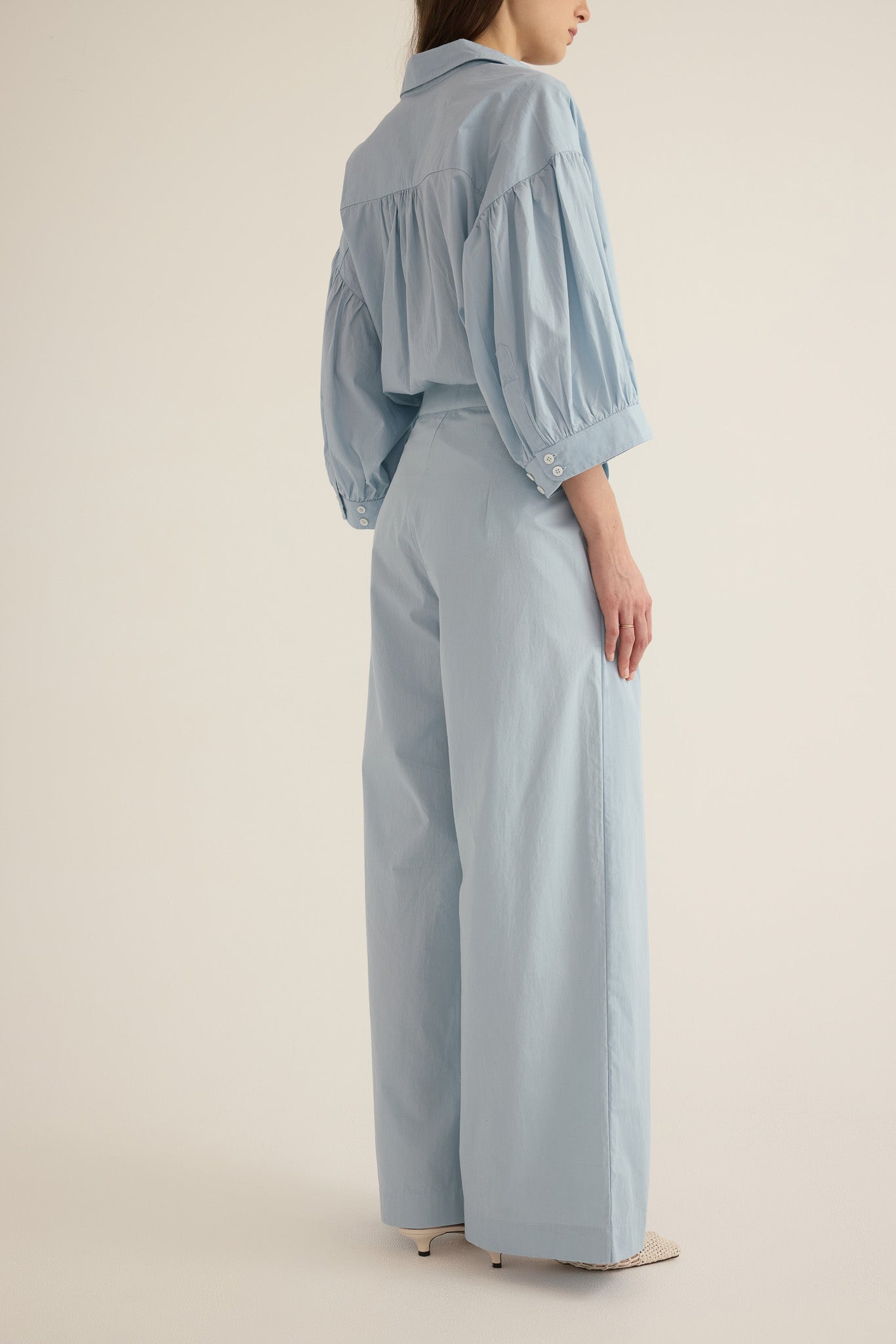 Side view of model wearing the Romeo wide-leg pleat pant in pale blue organic cotton, styled with the Romeo wide cuff blouson shirt in blue